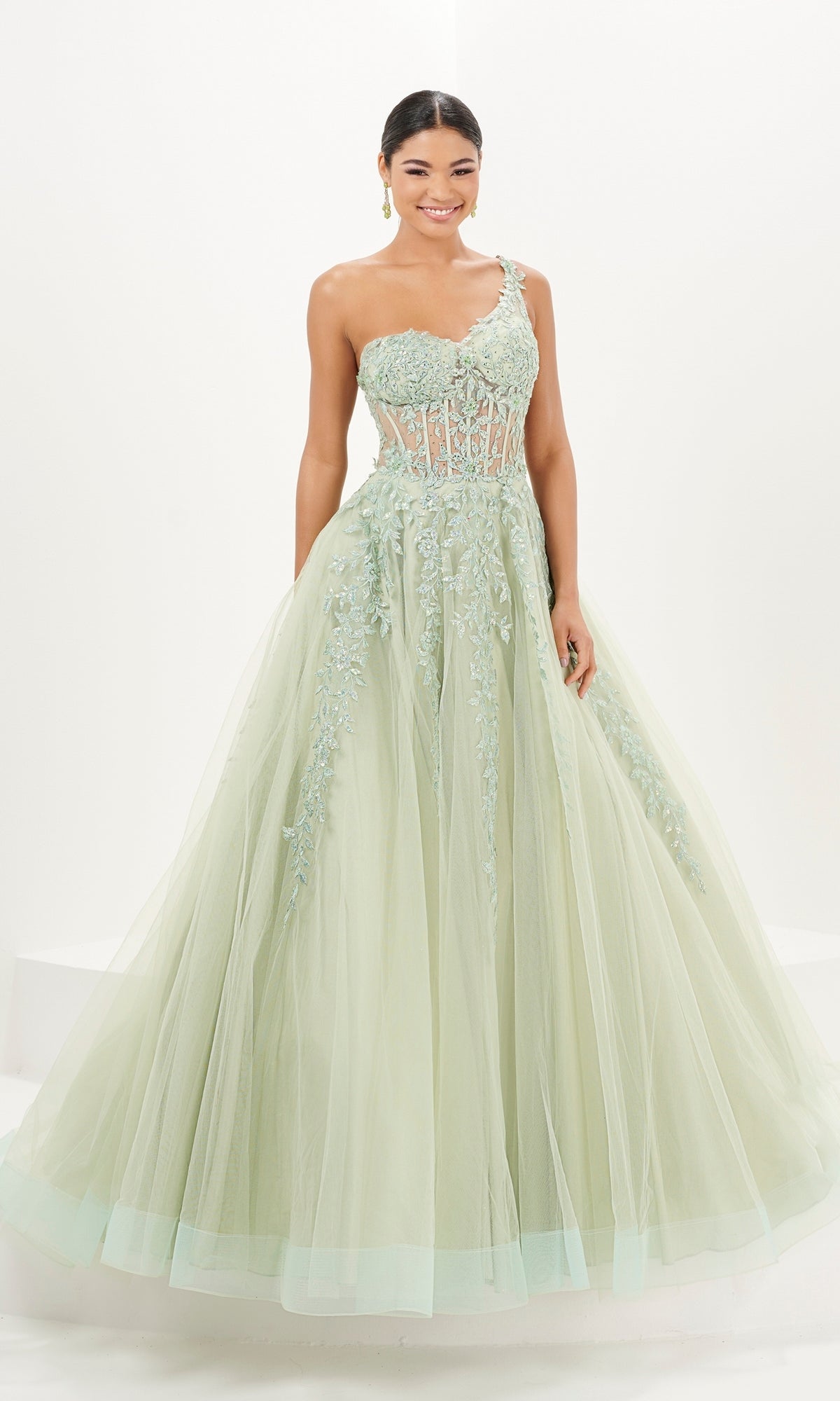 A woman wearing the Tiffany One-Shoulder Sheer Corset Prom Dress 16096, an a-line light green gown with floral appliques and a tulle skirt, stands smiling against a white background.